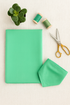 A neatly folded piece of Bella Solids Betty’s Green Fabric by Moda Fabrics lies on a beige surface beside gold sewing scissors, two wooden spools of green thread, and a small potted succulent under soft natural light.