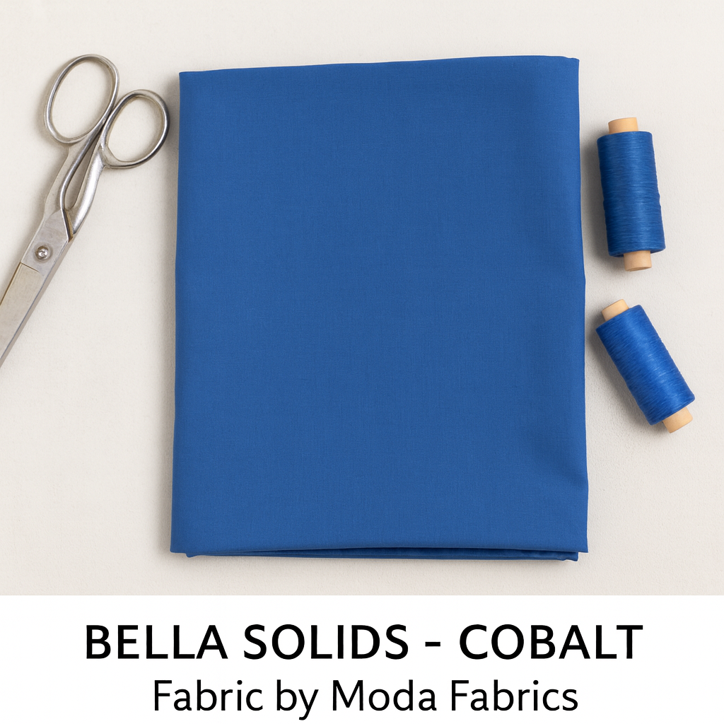 A folded piece of deep blue Bella Solids Cobalt Fabric by Moda Fabrics lies on a soft off-white surface beside silver sewing scissors and blue thread spools, captured under natural lighting with crisp, clean composition.