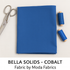 A folded piece of deep blue Bella Solids Cobalt Fabric by Moda Fabrics lies on a soft off-white surface beside silver sewing scissors and blue thread spools, captured under natural lighting with crisp, clean composition.