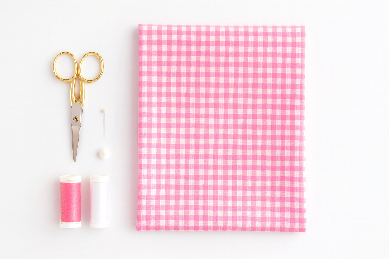 folded pink gingham fabric with thread spools, pins, and scissors on a white table.