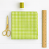 folded green gingham fabric with thread, scissors, and ruler on a white table.