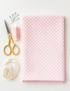 folded pink gingham-dot fabric with scissors, pink thread, pins, and lace ribbon on a white table.