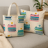 A cozy living room setting with a beige couch featuring a tote bag and pillow made from Ruby Star Society’s Best Day Ever Typewriters Natural Canvas fabric by Melody Miller. The design shows colorful retro typewriters with hearts in teal, pink, and green tones on natural cotton-linen canvas.