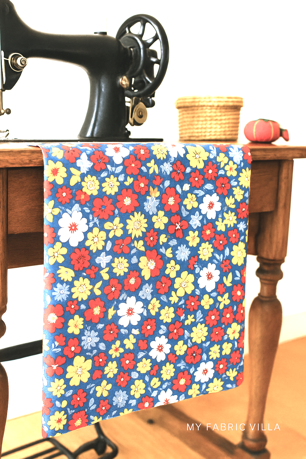 A cozy sewing room setup featuring a vintage patchwork quilt and colorful fabric swatches made from 30s Playtime 2025 Bouncy Florals Bluebell fabric by Linzee McCray for Moda Fabrics, with cheerful red, yellow, white, and blue flowers on a blue background and a “My Fabric Villa” watermark in the bottom-right corner.