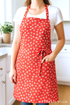 A woman stands in a bright sewing studio wearing a vintage-style apron made from red floral fabric featuring white blossoms and pink apples. The apron is crafted from 30s Playtime 2025 Apple Blossom Florals Scarlet Fabric by Linzee McCray for Moda Fabrics, with a “My Fabric Villa” watermark in the bottom-right corner.