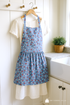 A vintage-style apron made from 30s Playtime 2025 - Apple Blossom Florals Sky Fabric by Linzee McCray for Moda Fabrics hangs on a hook in a cozy farmhouse kitchen. The soft blue cotton fabric features red apples and white blossoms, illuminated by natural light with wooden countertops and pastel dishware in the background. A subtle “My Fabric Villa” watermark appears in the bottom-right corner.