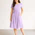 A woman wearing a vintage-style lilac dress made from 30s Playtime 2025 Perky Posies Lilac Fabric by Linzee McCray for Moda Fabrics, featuring white floral prints and a flared skirt, displayed in a softly lit sewing studio.