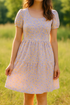 A woman standing outdoors in a sunny field wearing a light purple and yellow floral cotton dress with short puff sleeves and a gathered waist, made from vintage-inspired 1930s floral fabric.