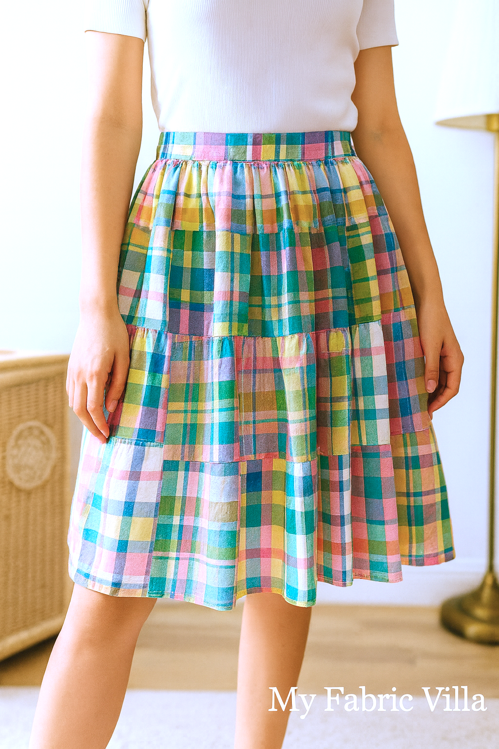 A close-up photograph of a woman indoors wearing a knee-length pastel madras plaid skirt made from the Nantucket Patchwork Sorbet fabric, featuring soft pink, blue, yellow, and green tones; the image includes a “My Fabric Villa” watermark in the bottom right corner.