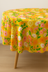 A close-up of a round dining table covered with a vibrant tablecloth made from Best Day Ever – Foxgloves Florals Butterflies Golden Hour Fabric by Melody Miller for Ruby Star Society, featuring pink, orange, and white flowers with butterflies on a golden-yellow background in a bright, modern dining room.