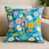 A decorative square pillow made from the Best Day Ever – Foxgloves Florals Butterflies Summer Sky Fabric by Melody Miller for Ruby Star Society, featuring teal-blue background with colorful orange, pink, yellow, and white flowers and butterflies, displayed on a light neutral sofa