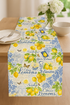 A bright dining table decorated with a cotton table runner made from Limoncello Lemon Collage Fabric by Timeless Treasures, featuring yellow lemons, blue butterflies, and cursive text on an ivory background with blue damask accents, illuminated by soft white indoor lighting.