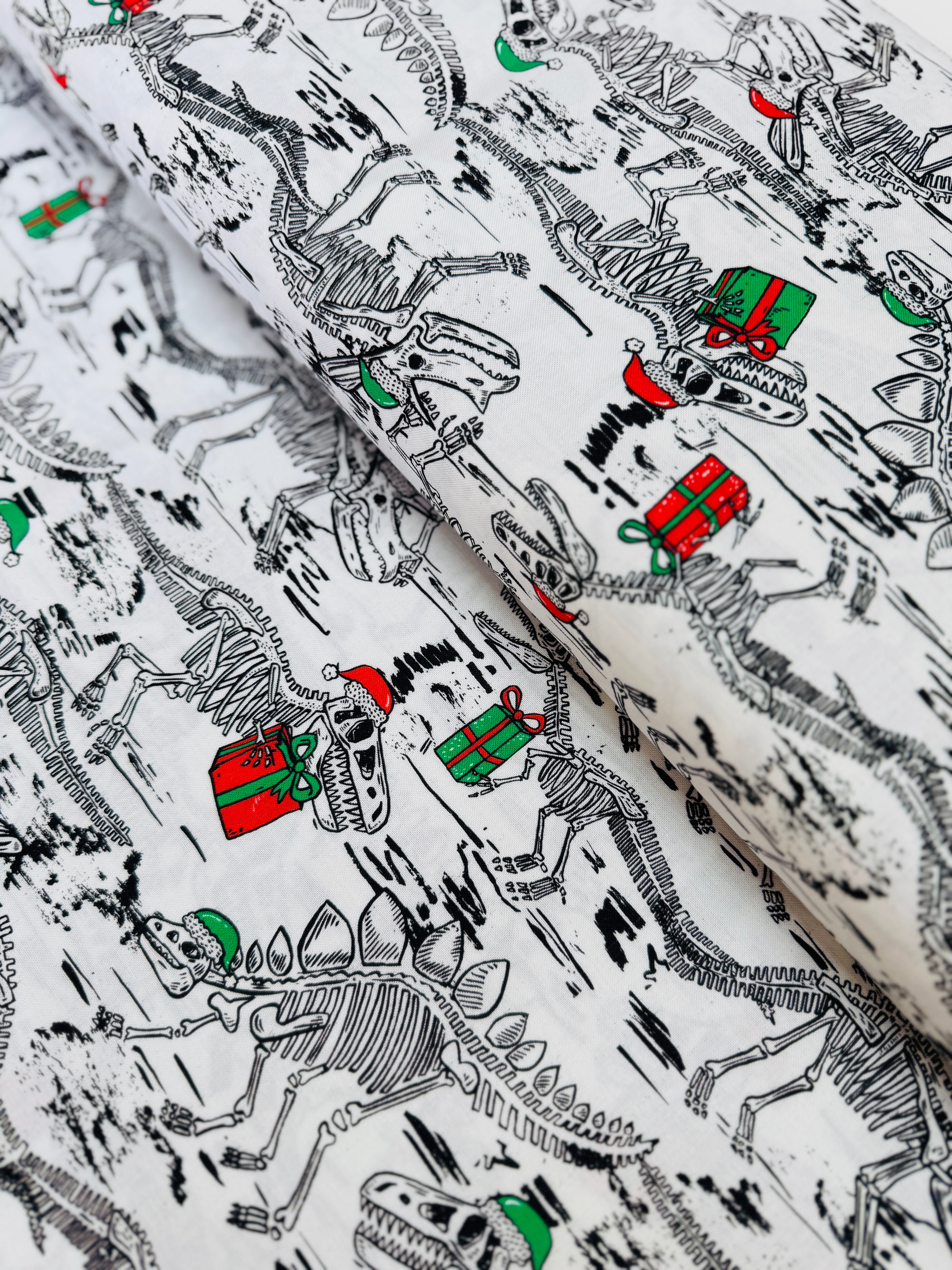 White cotton fabric with skeleton dinosaur T‑Rex prints wearing Santa hats and holding presents