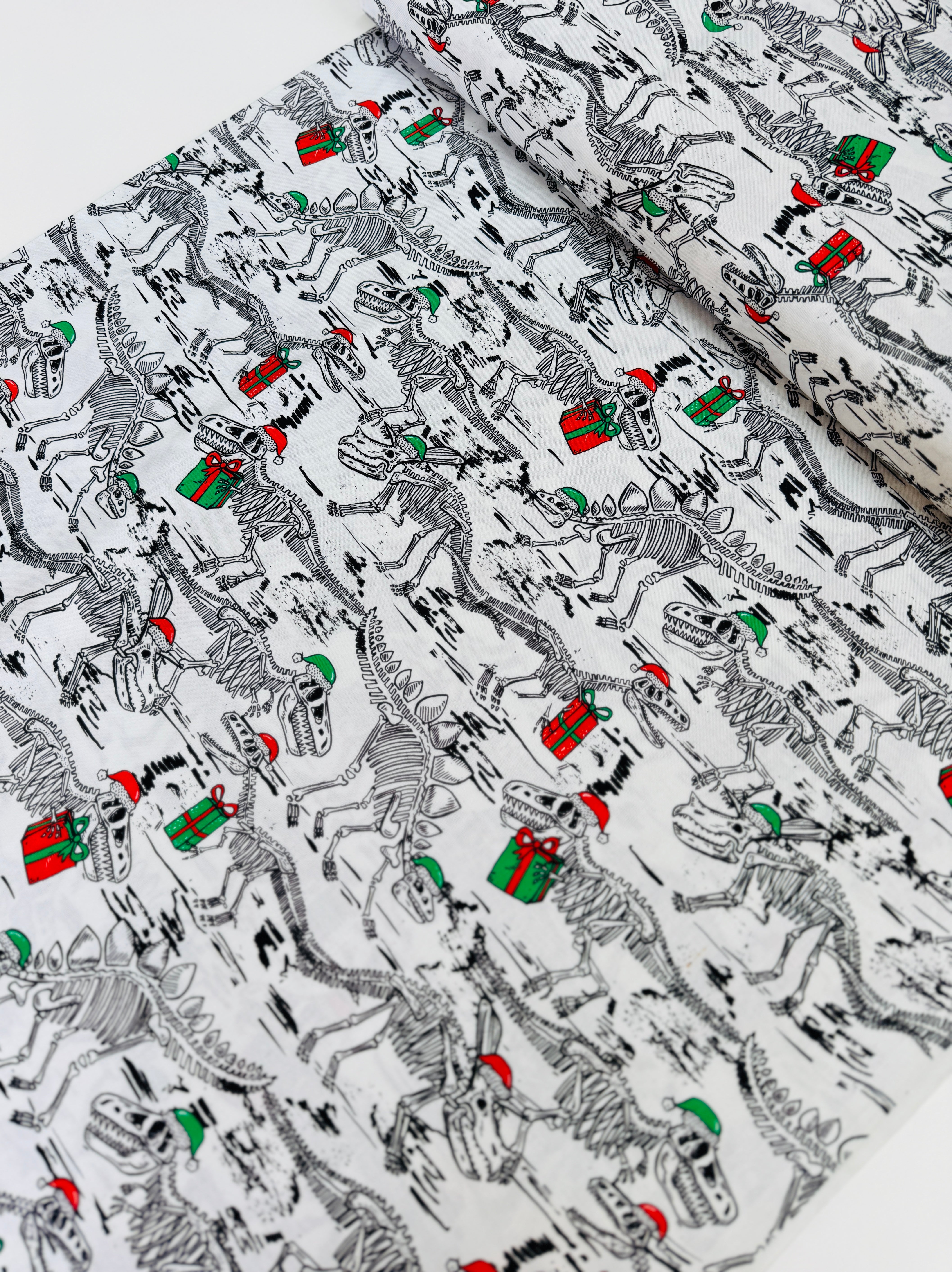 White cotton fabric with skeleton dinosaur T‑Rex prints wearing Santa hats and holding presents