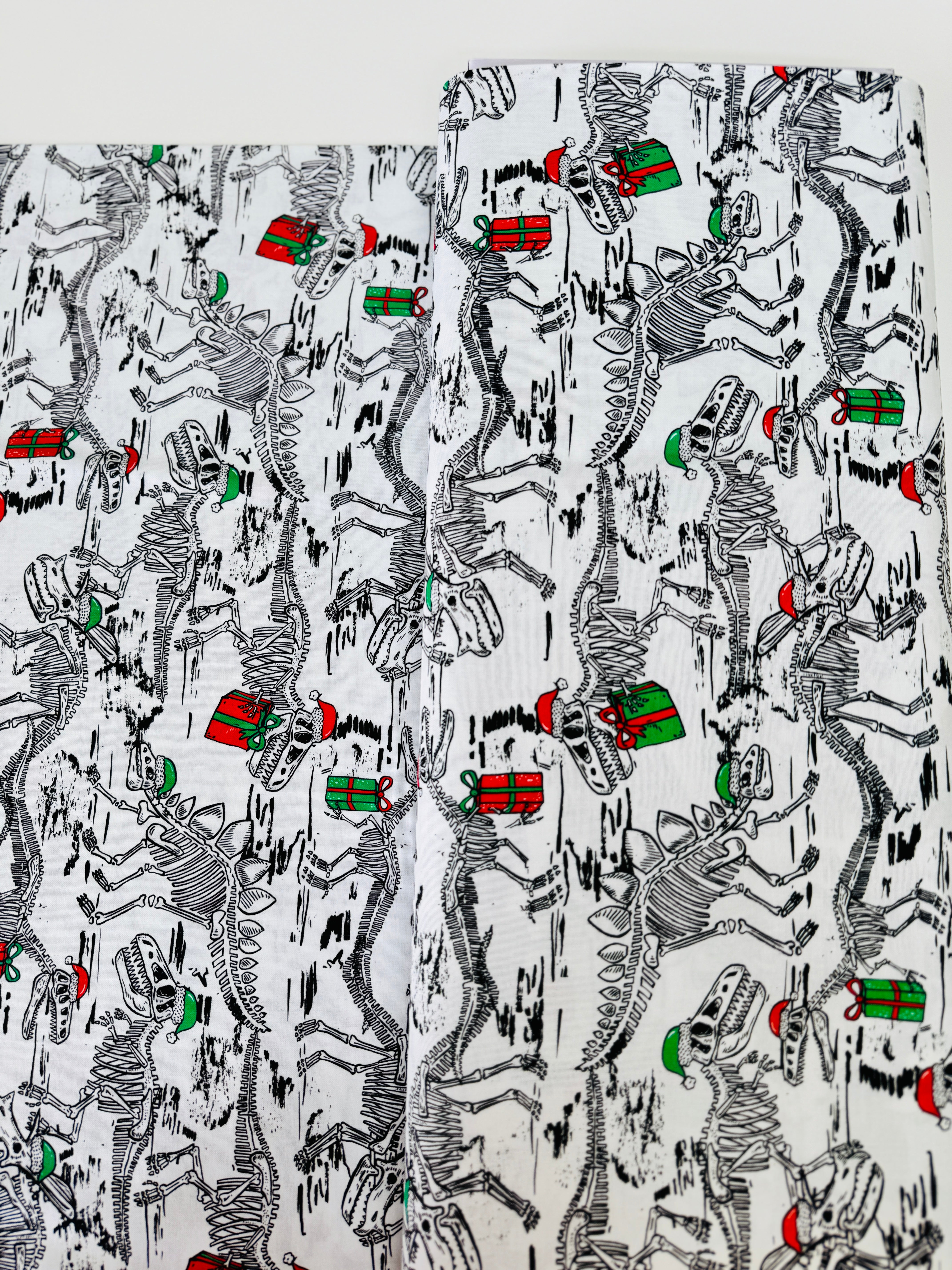 White cotton fabric with skeleton dinosaur T‑Rex prints wearing Santa hats and holding presents