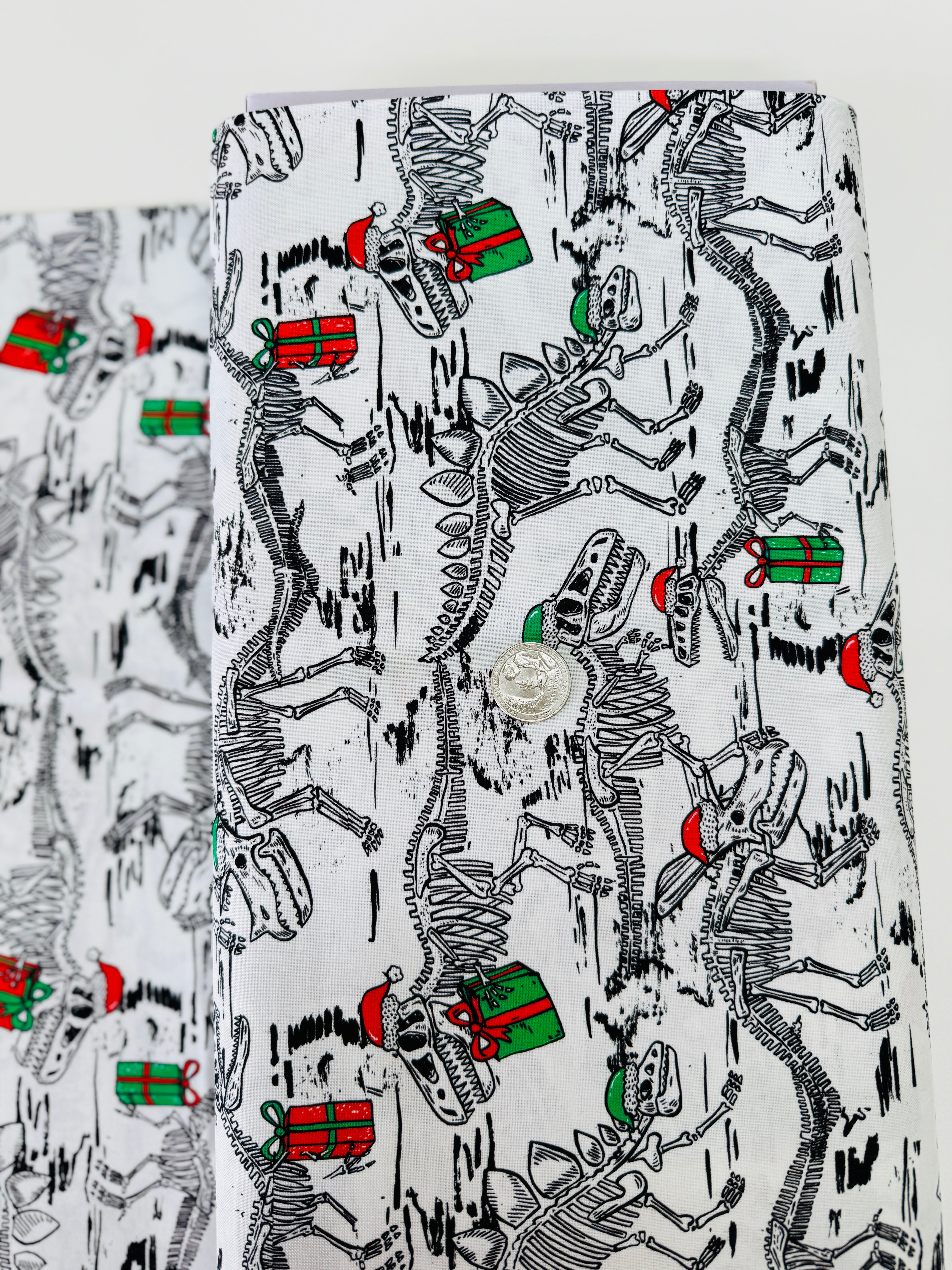 White cotton fabric with skeleton dinosaur T‑Rex prints wearing Santa hats and holding presents