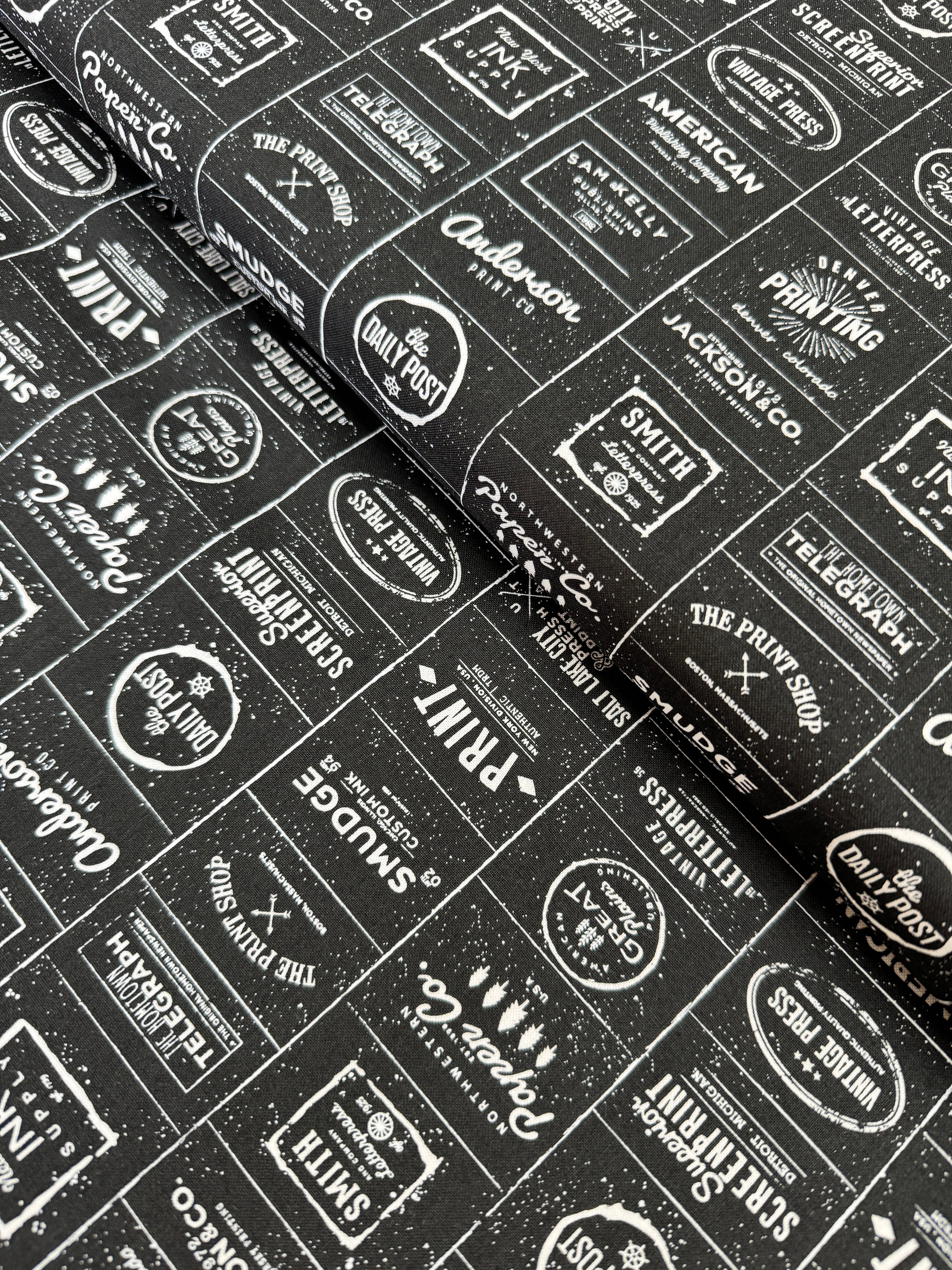 The Print Shop Modern Logos Black fabric by Sweetwater for Moda Fabrics featuring white vintage typography logos on a black background.