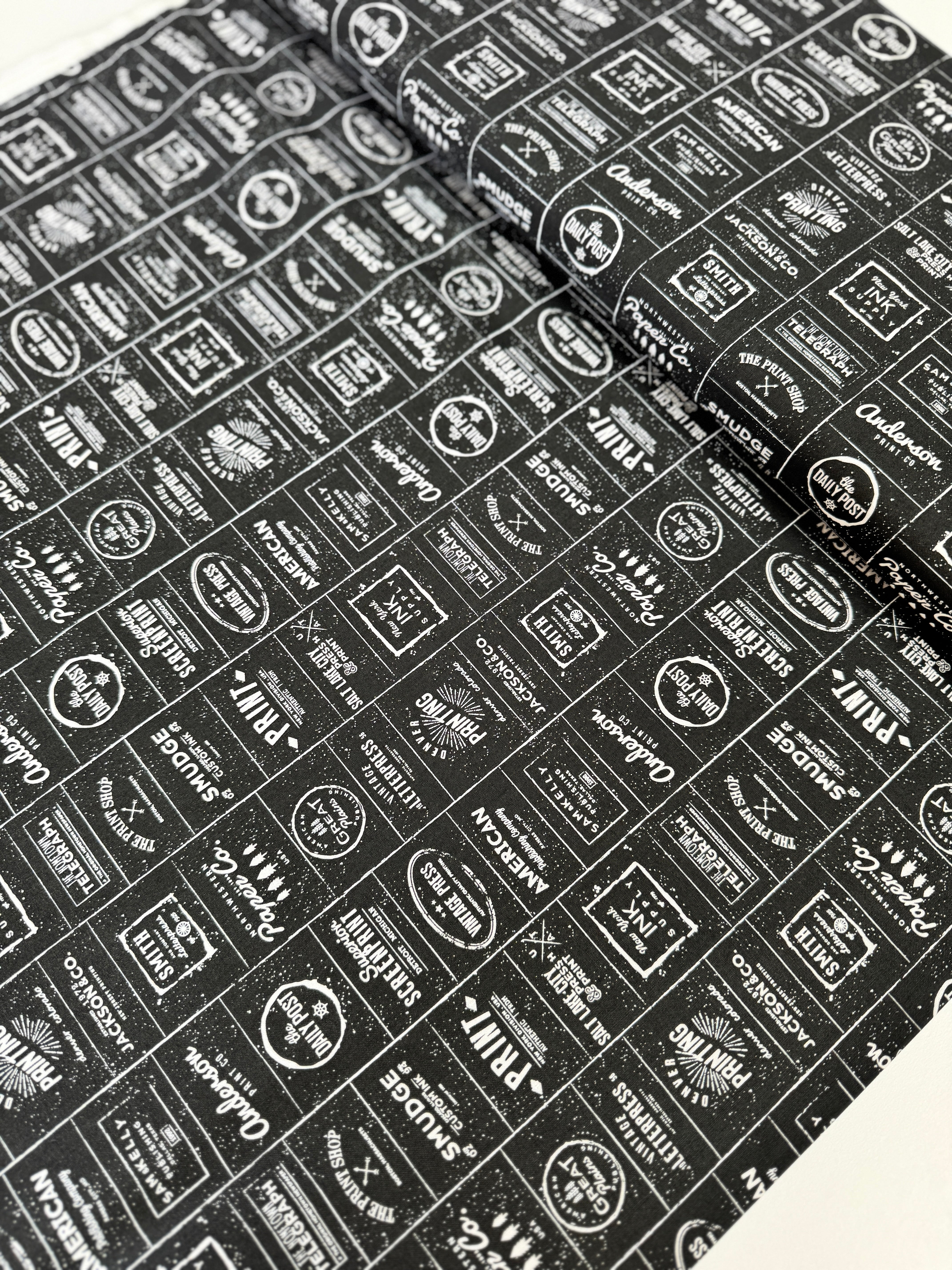 The Print Shop Modern Logos Black fabric by Sweetwater for Moda Fabrics featuring white vintage typography logos on a black background.