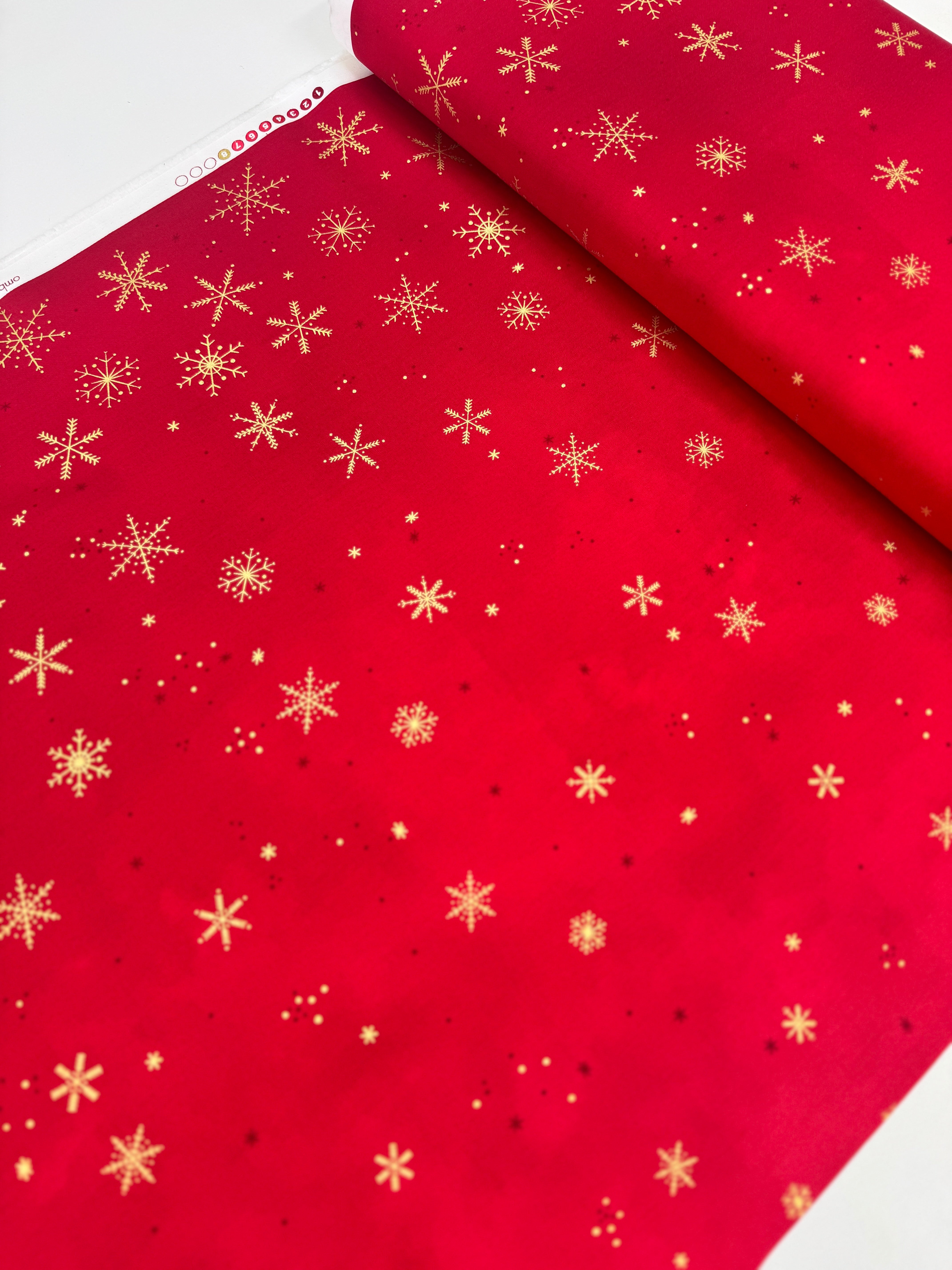 A vibrant red fabric from the Ombre Flurries Metallic collection by V and Co. for Moda Fabrics, featuring shimmering gold metallic snowflakes scattered across the surface.