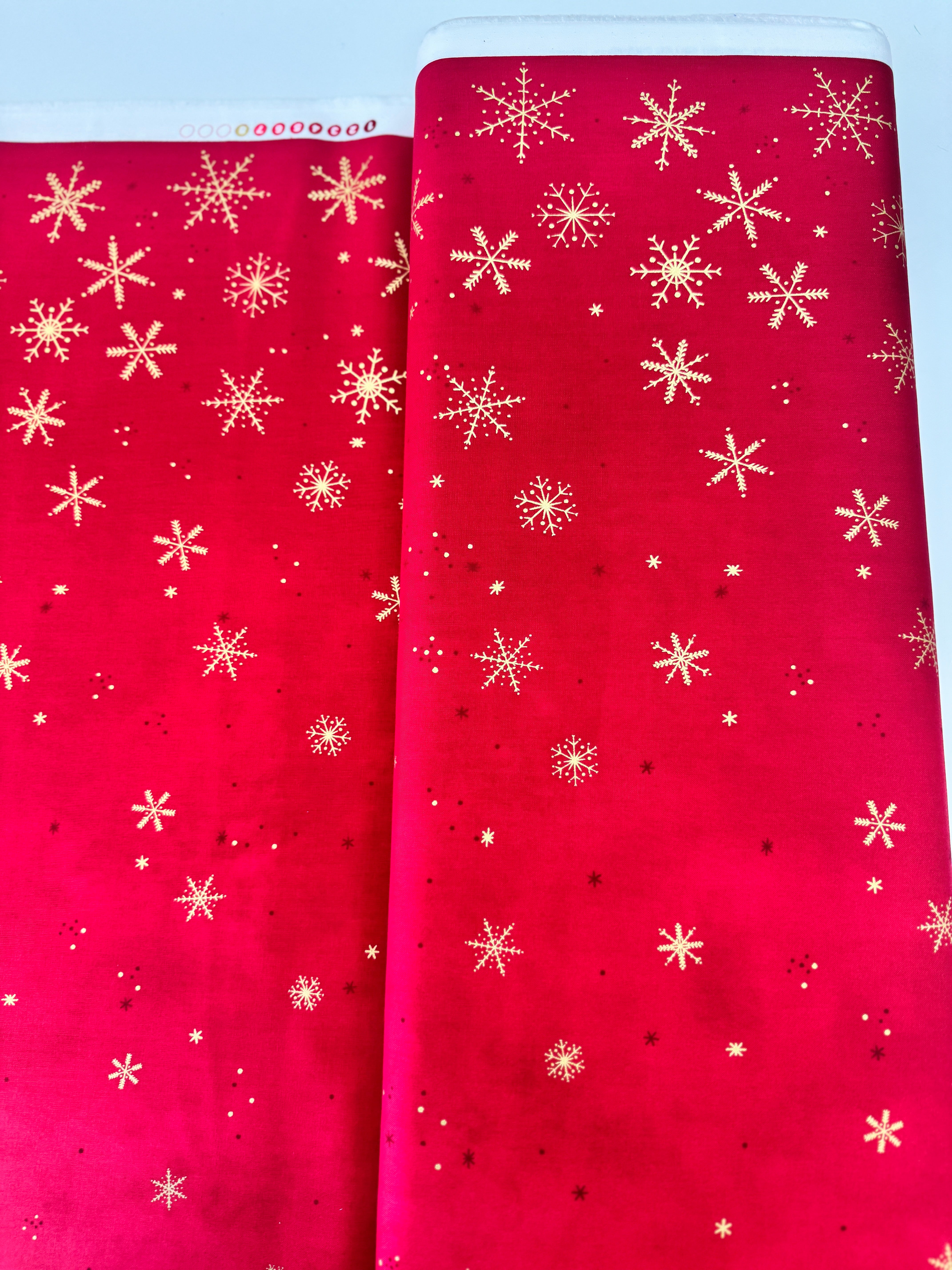 A vibrant red fabric from the Ombre Flurries Metallic collection by V and Co. for Moda Fabrics, featuring shimmering gold metallic snowflakes scattered across the surface.