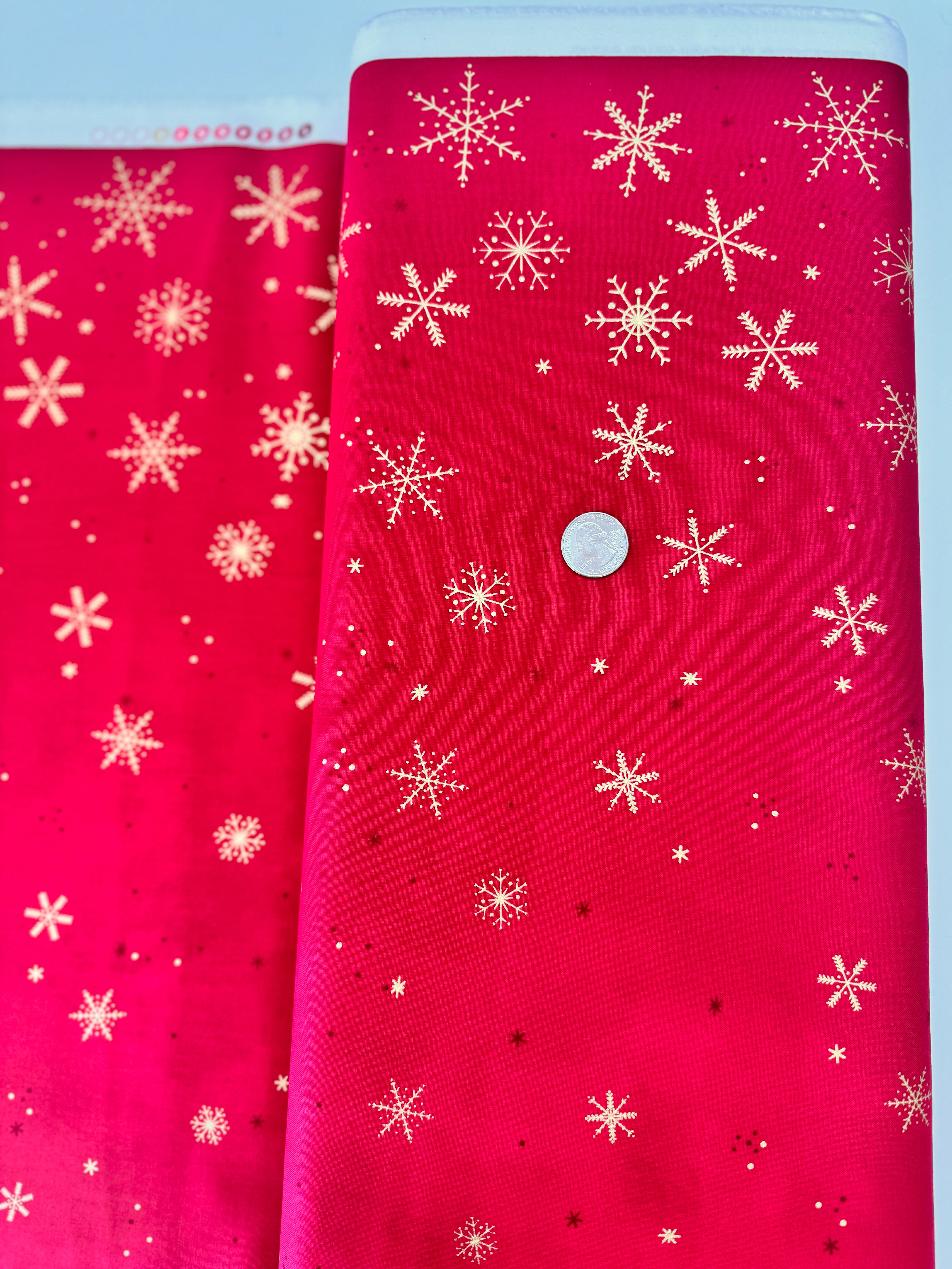 A vibrant red fabric from the Ombre Flurries Metallic collection by V and Co. for Moda Fabrics, featuring shimmering gold metallic snowflakes scattered across the surface.