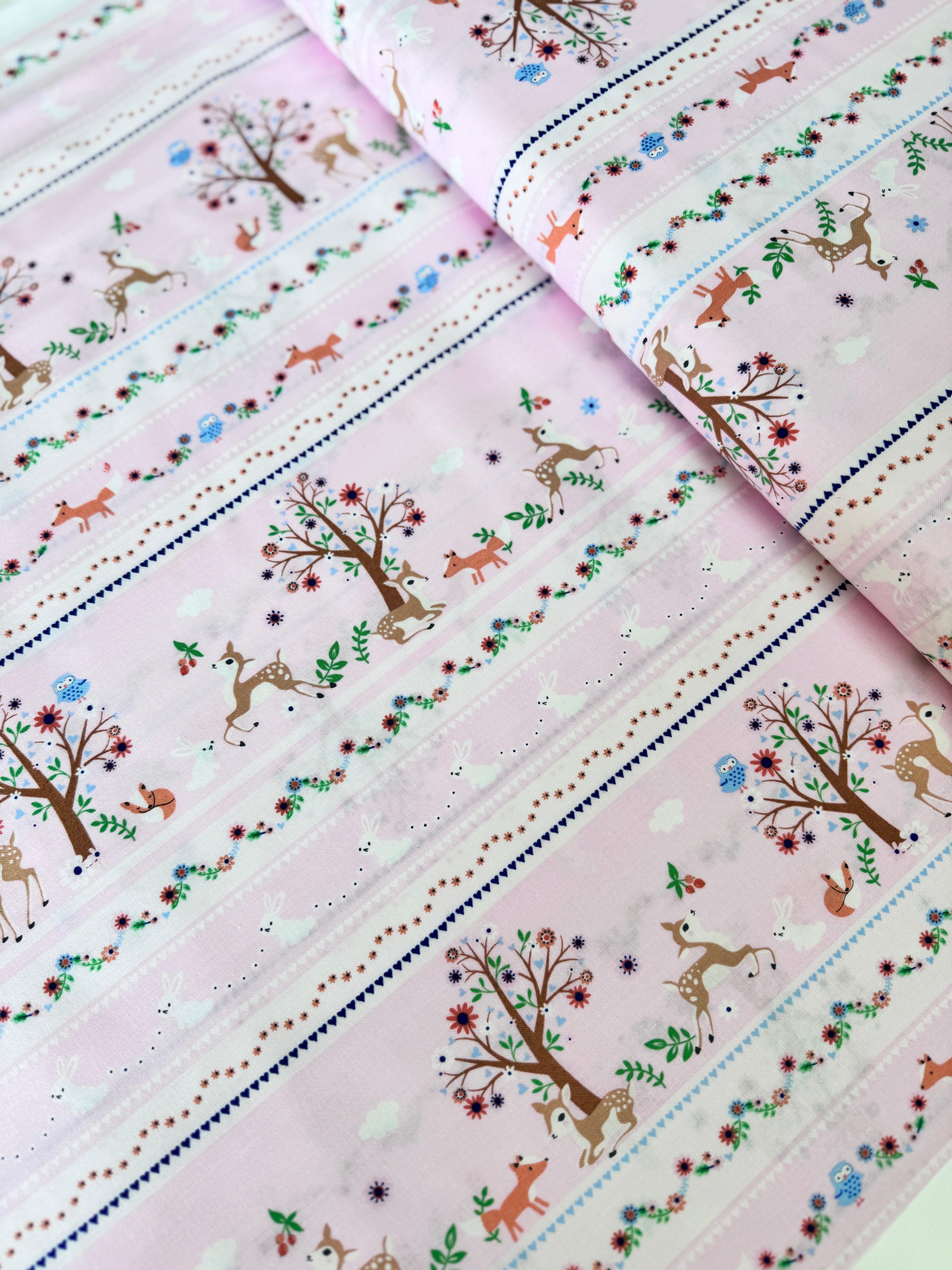 A pastel pink cotton fabric featuring rows of playful deer, white bunnies, trees, and flowers in a stripe pattern - Storybook Forest - Bunnies & Deer Stripe by Timeless Treasures.