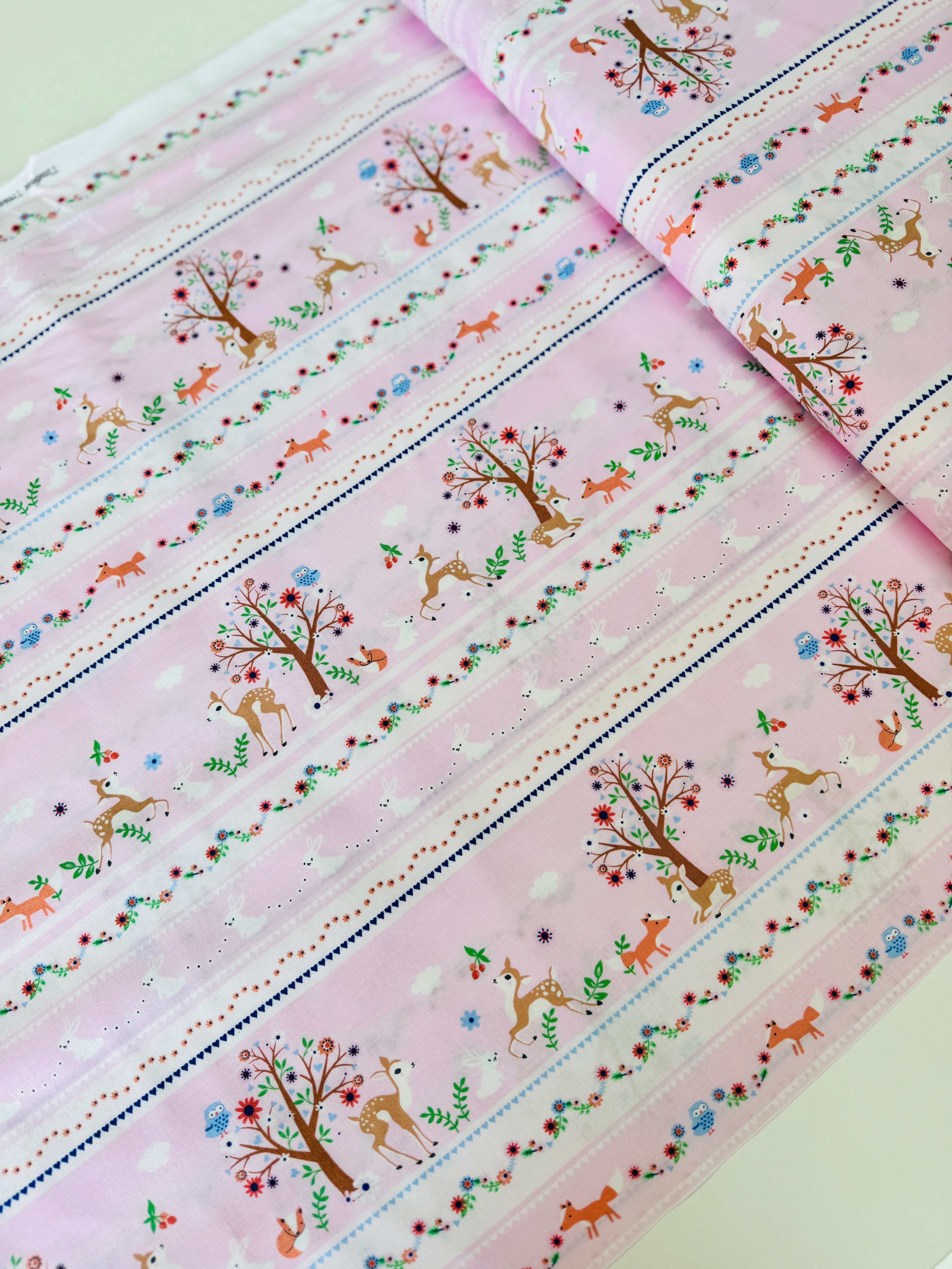 A pastel pink cotton fabric featuring rows of playful deer, white bunnies, trees, and flowers in a stripe pattern - Storybook Forest - Bunnies & Deer Stripe by Timeless Treasures.