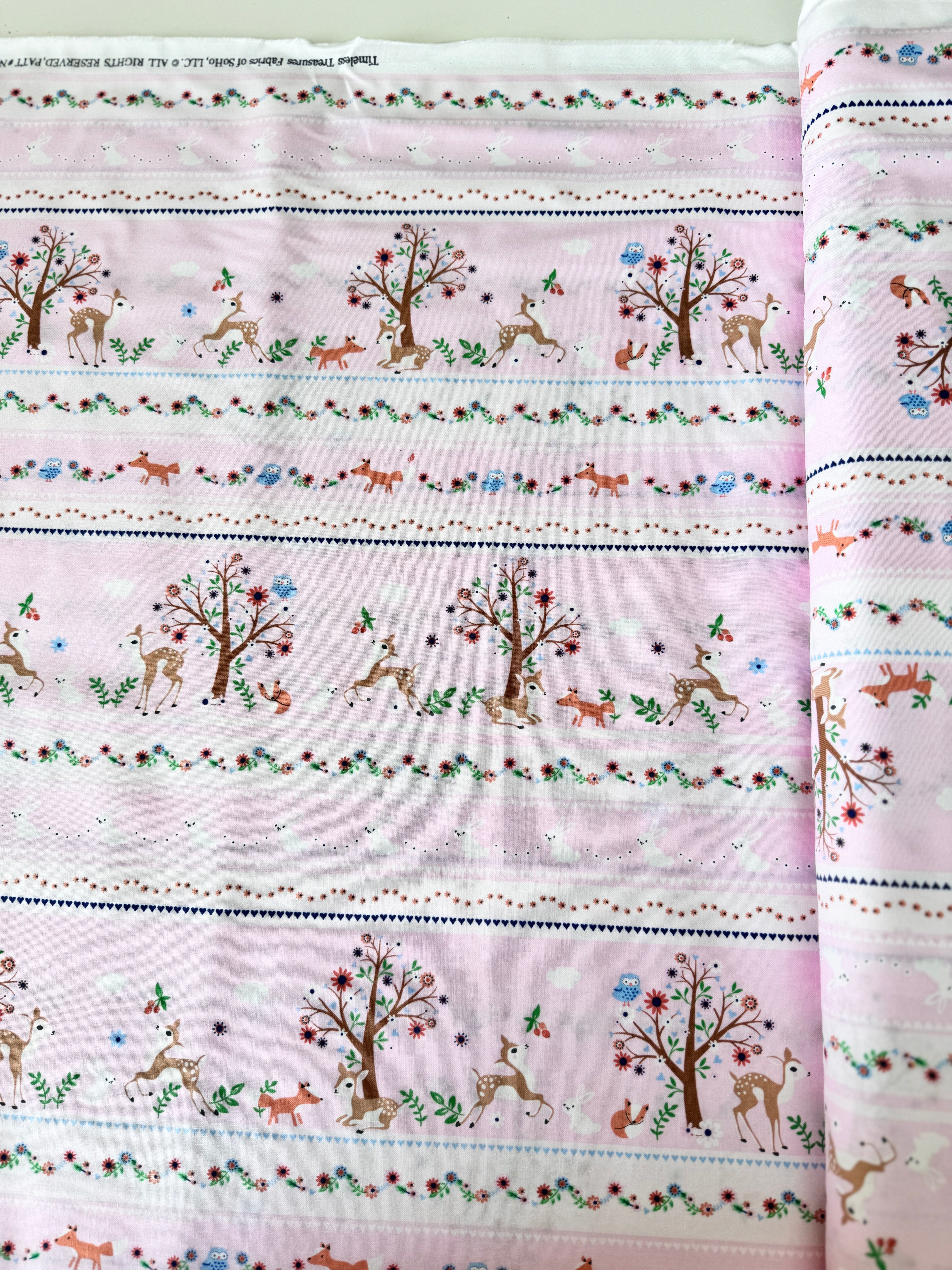 A pastel pink cotton fabric featuring rows of playful deer, white bunnies, trees, and flowers in a stripe pattern - Storybook Forest - Bunnies & Deer Stripe by Timeless Treasures.