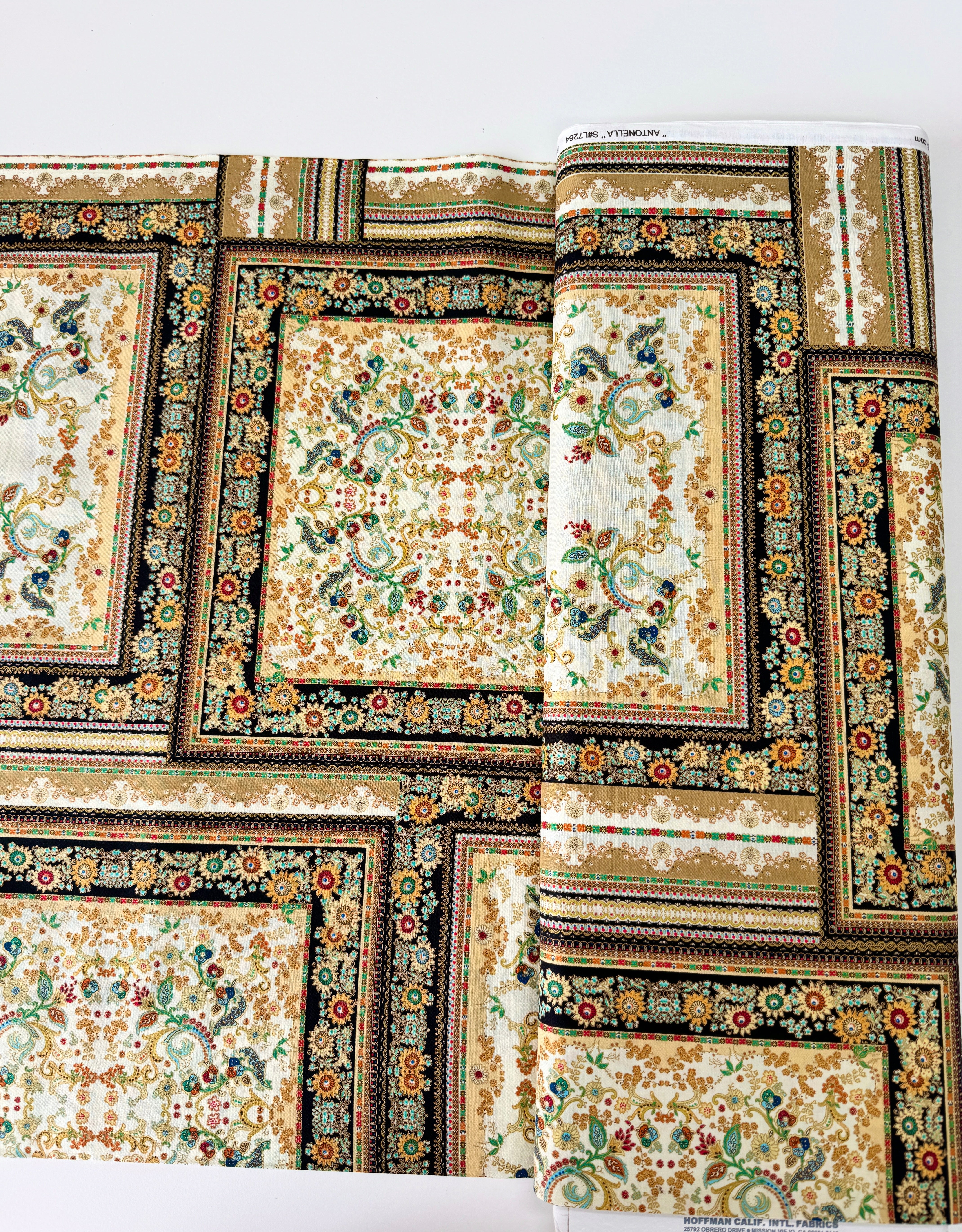 A close-up photo of Hoffman Fabrics’ Antonella Vintage Gold Metallic Fabric showing an ornate floral and scroll tapestry design in gold, cream, and black with shimmering metallic accents.