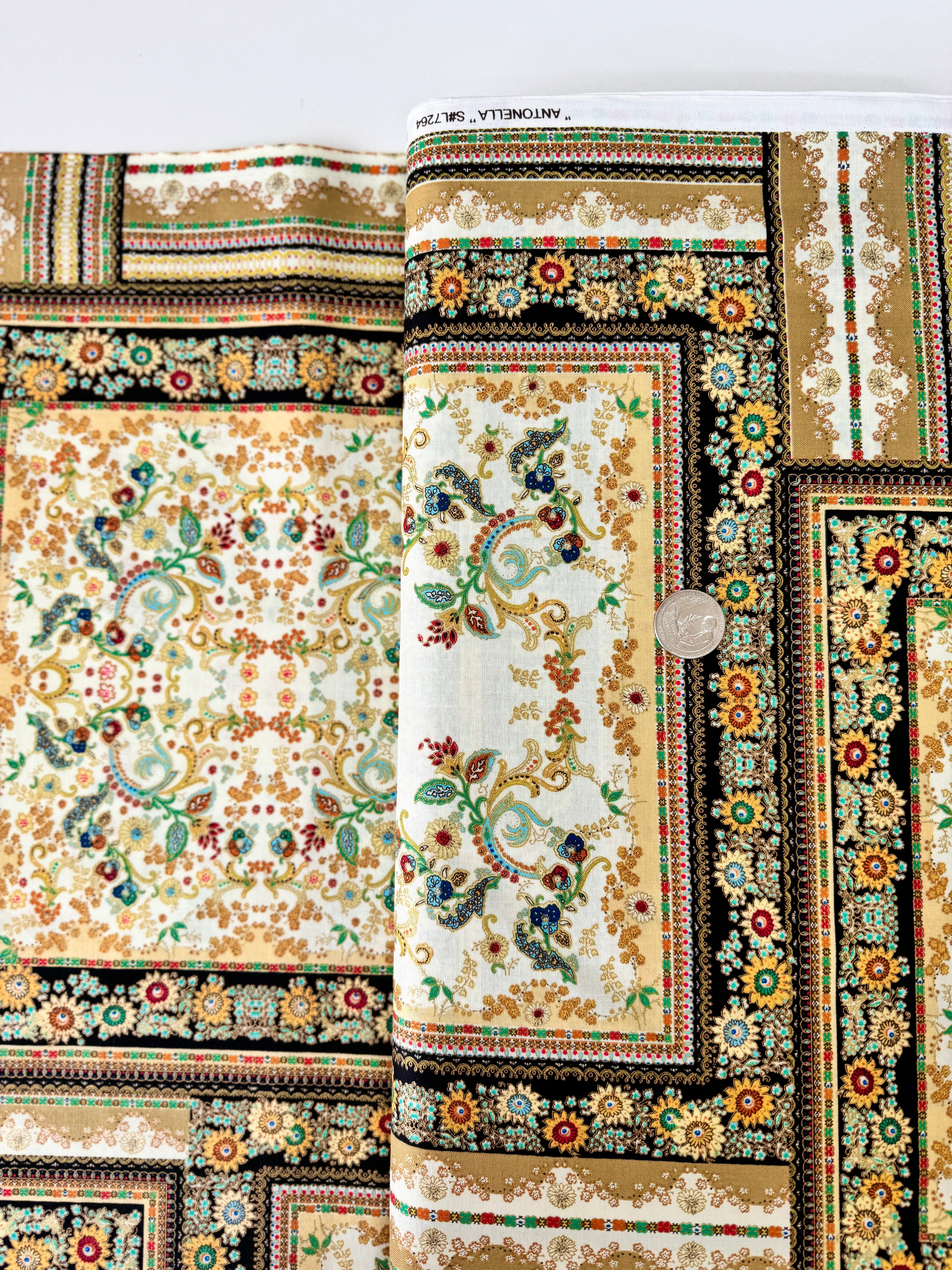 A close-up photo of Hoffman Fabrics’ Antonella Vintage Gold Metallic Fabric showing an ornate floral and scroll tapestry design in gold, cream, and black with shimmering metallic accents.