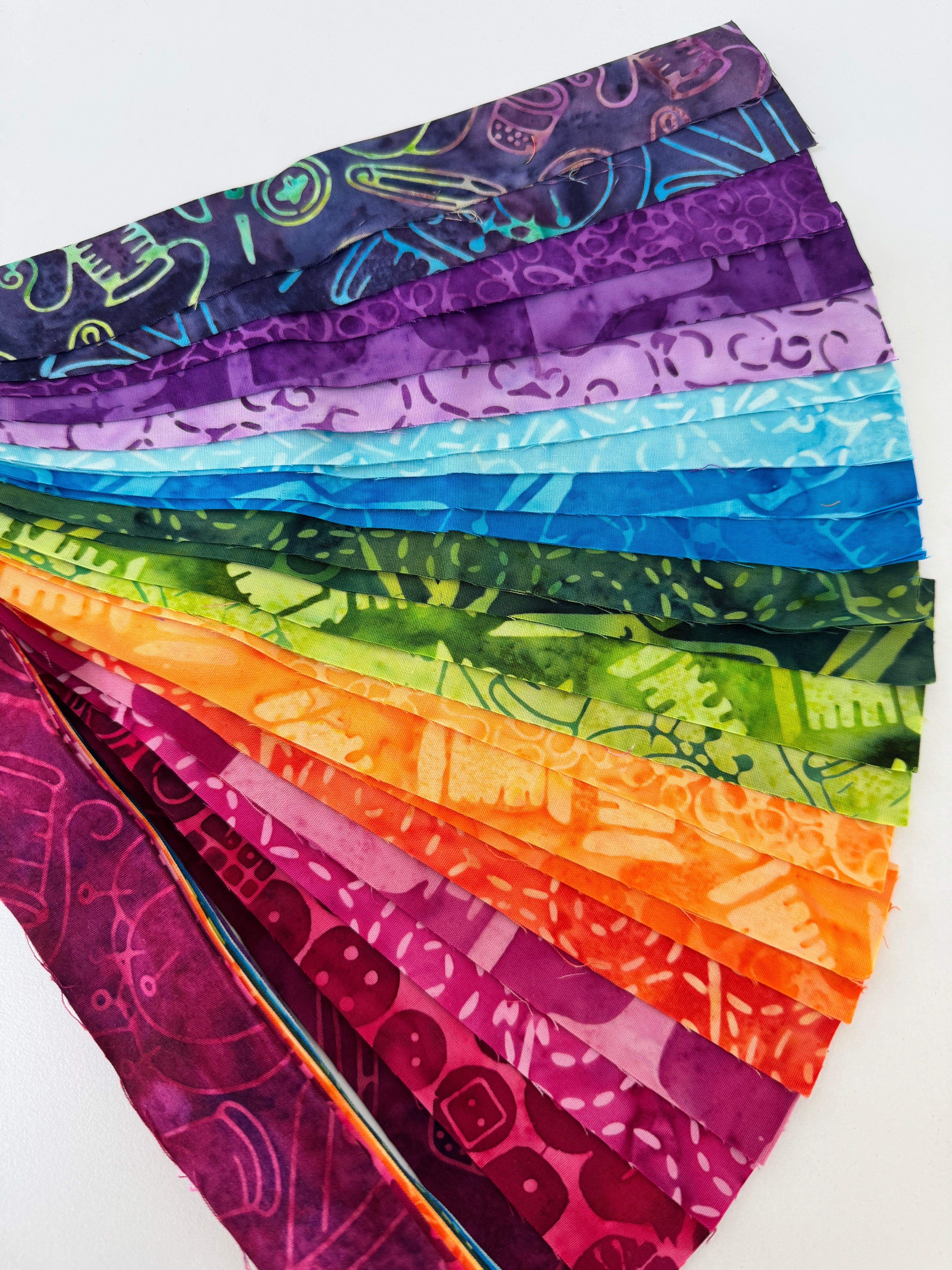 Assorted rainbow batik fabric strips fanned out, featuring 21 colorful 2-inch strips by Kathy Engle for Island Batik.