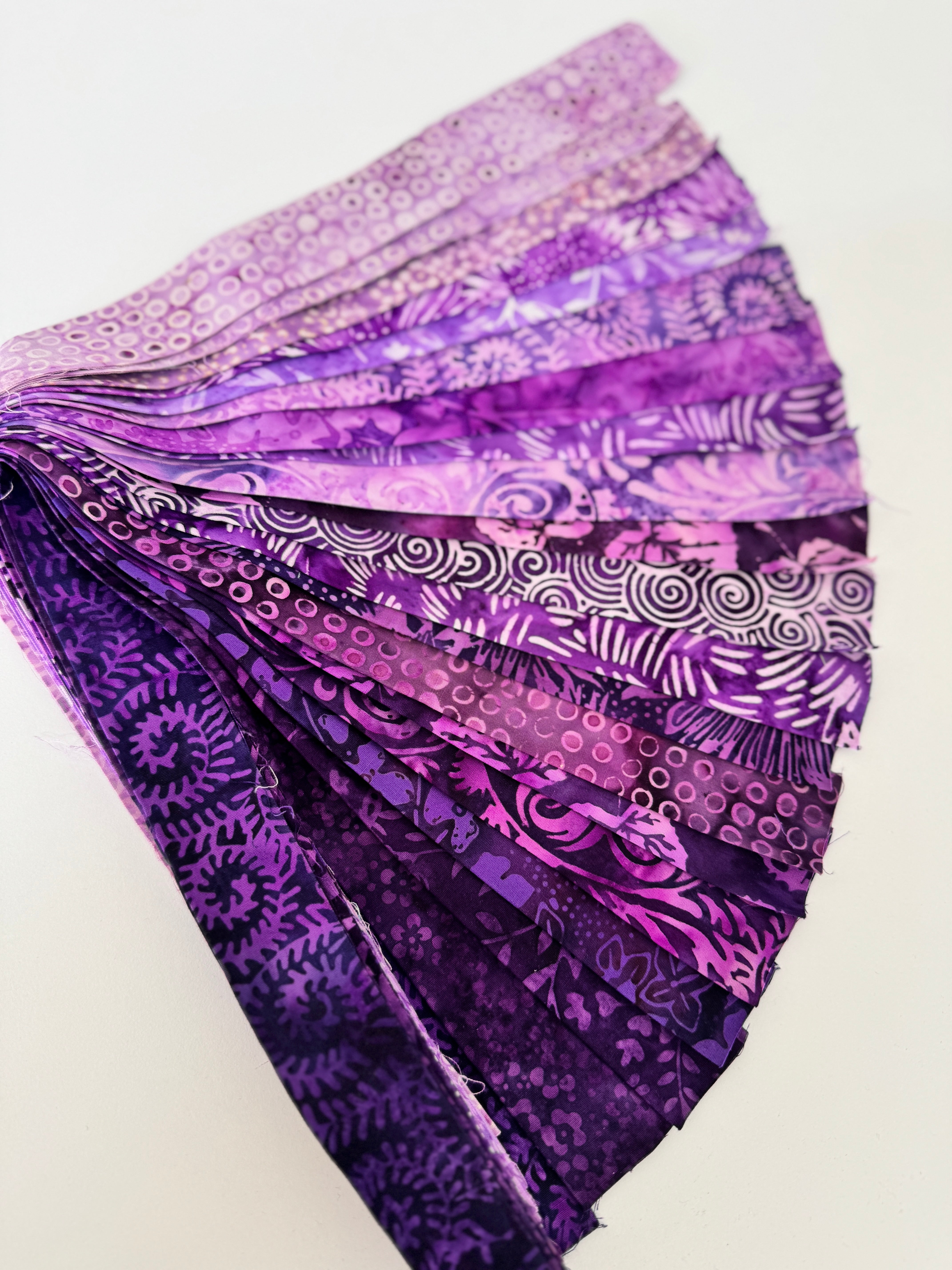 Assorted purple batik fabric strips fanned out, featuring 21 lavender to deep violet 2-inch strips by Kathy Engle for Island Batik.