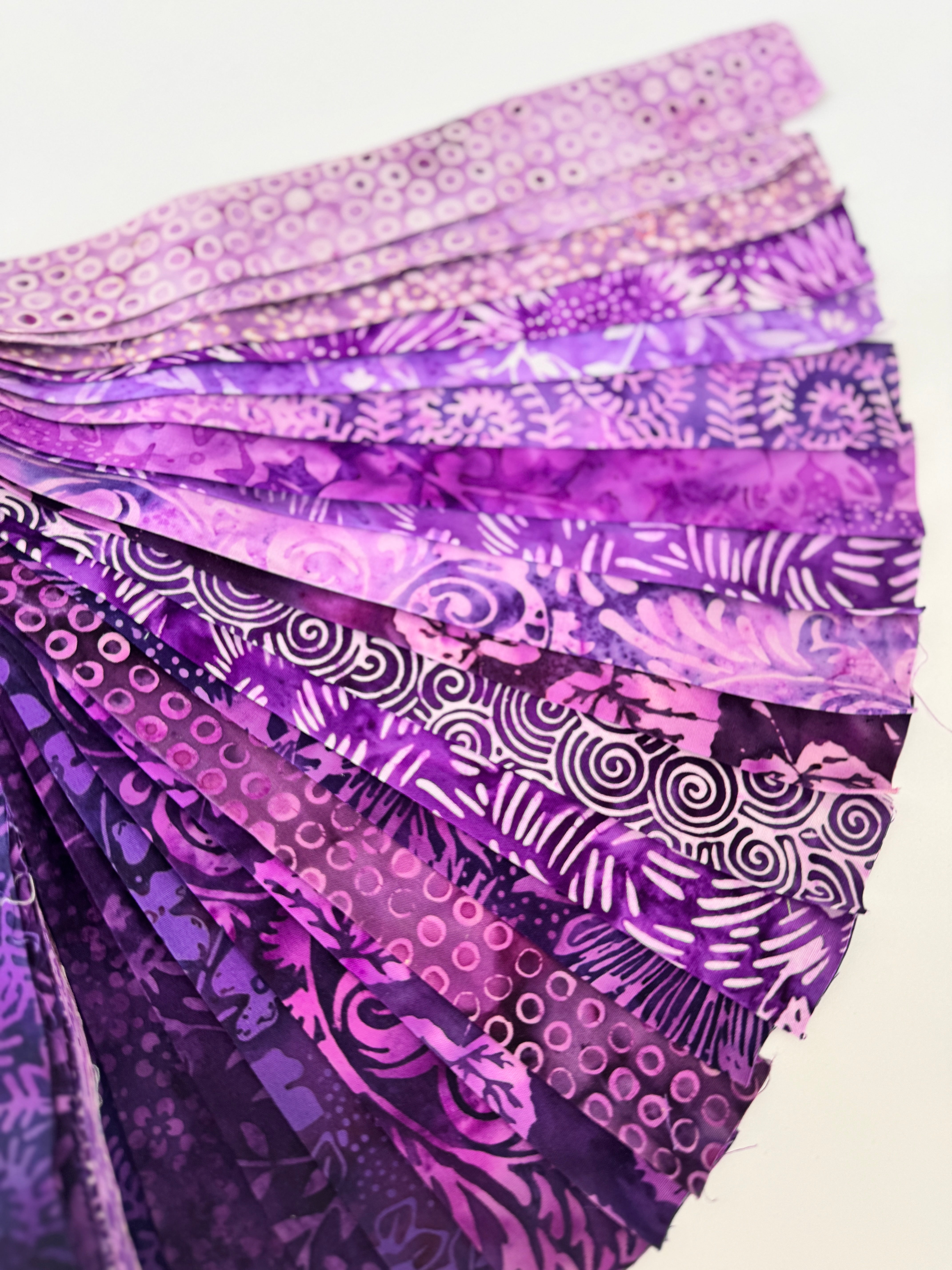 Assorted purple batik fabric strips fanned out, featuring 21 lavender to deep violet 2-inch strips by Kathy Engle for Island Batik.