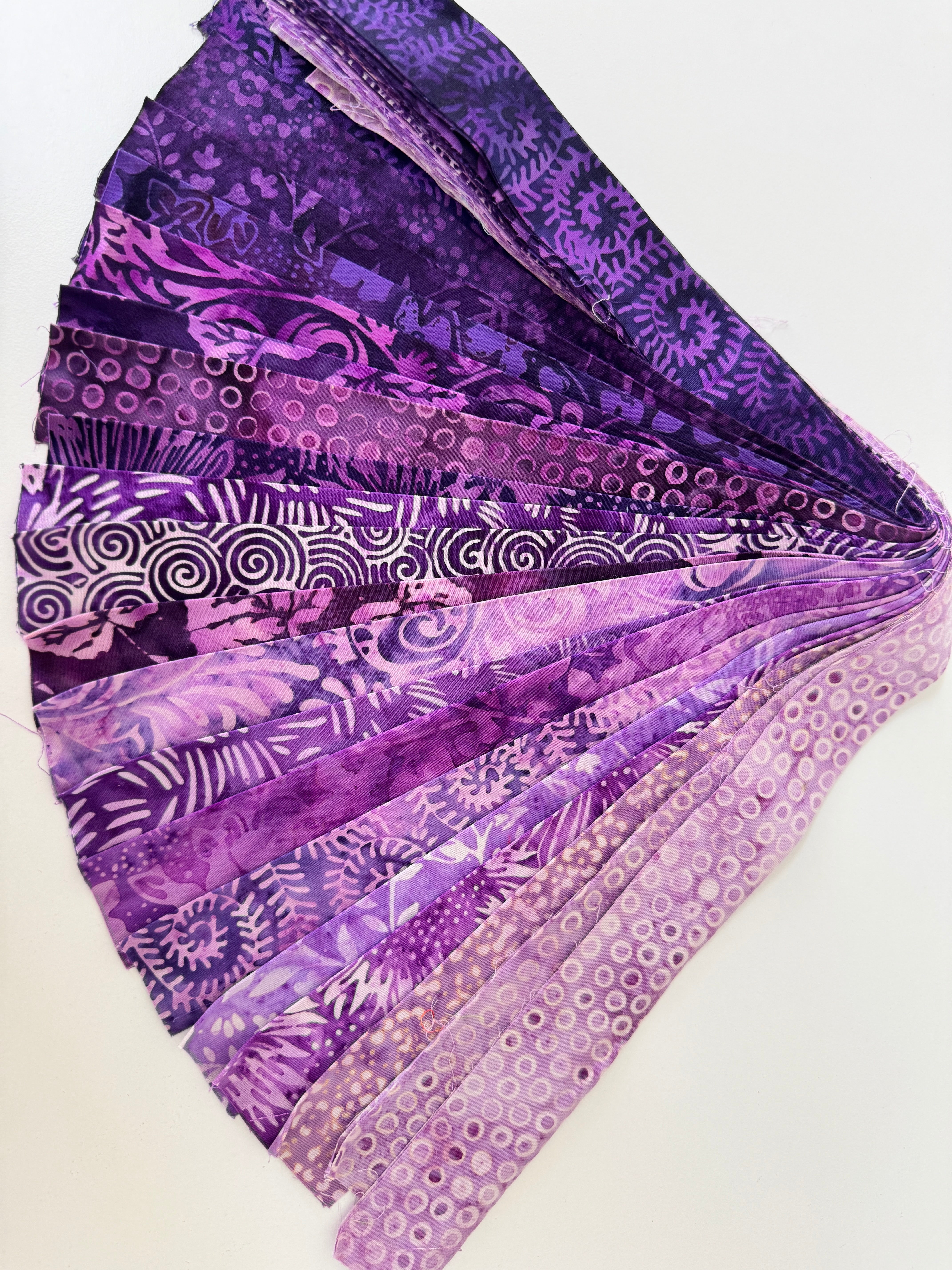 Assorted purple batik fabric strips fanned out, featuring 21 lavender to deep violet 2-inch strips by Kathy Engle for Island Batik.