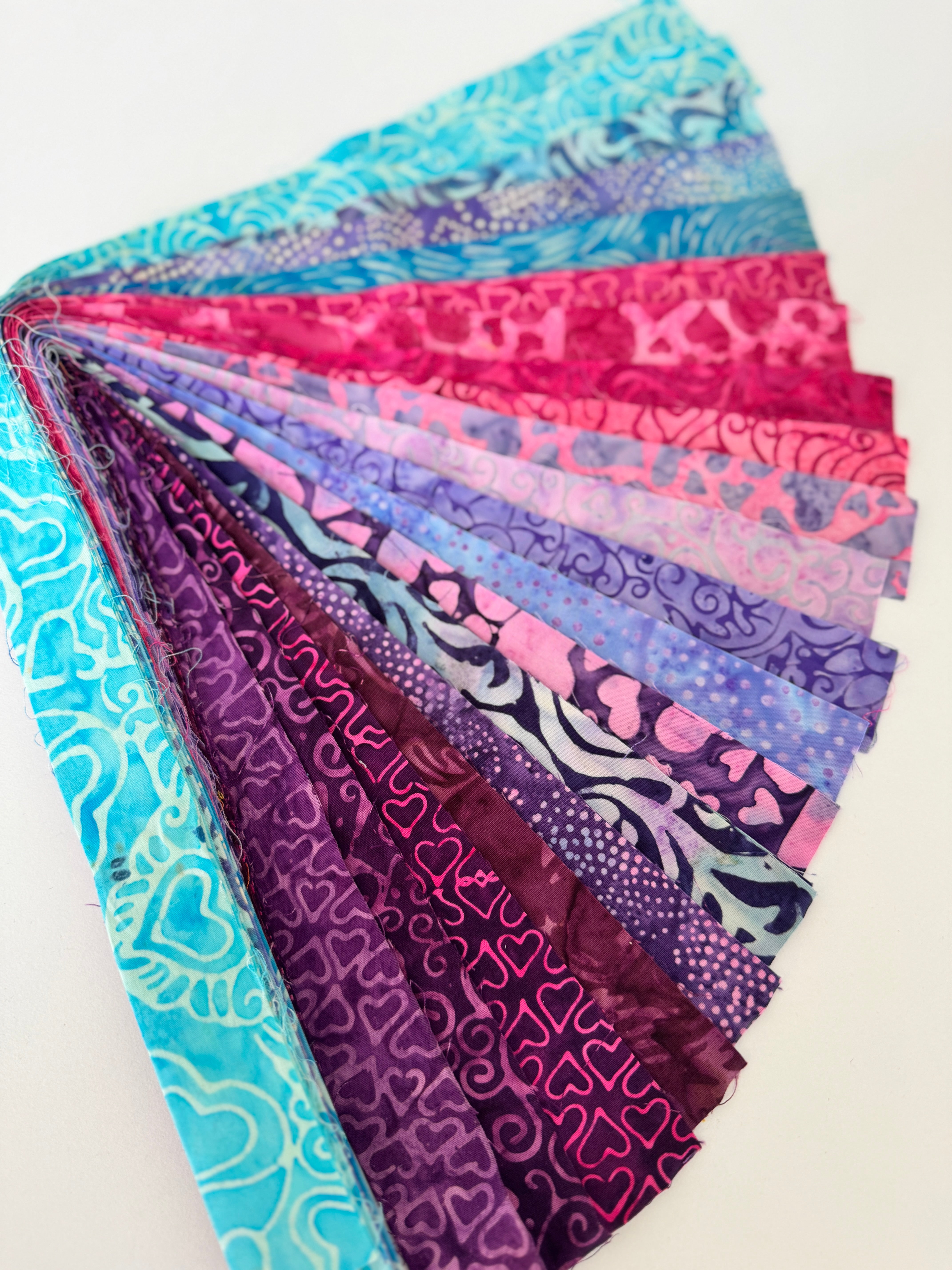 Bundle of assorted hearts-themed batik fabric strips in teal, pink, and purple tones, fanned out for display.