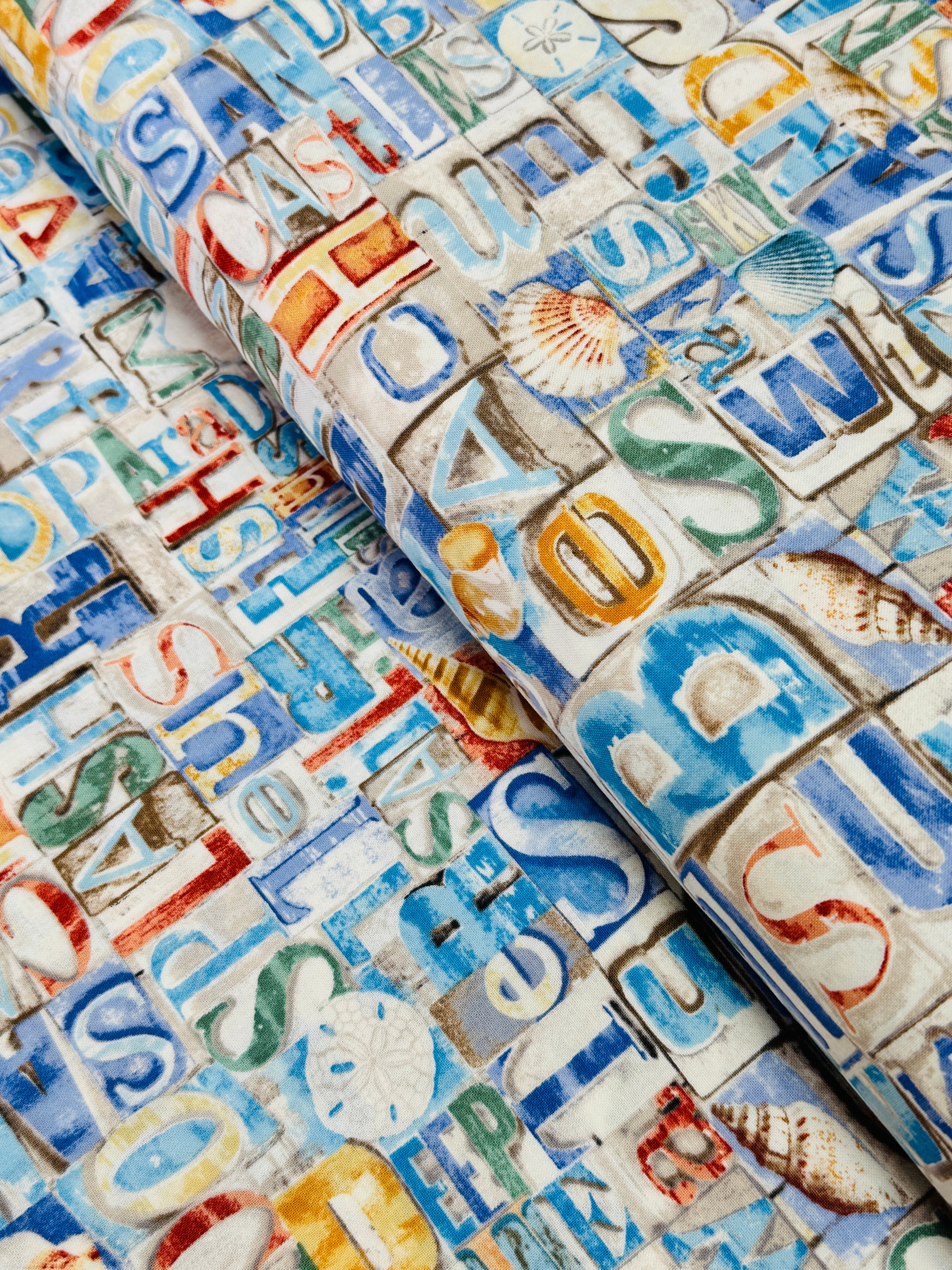 Coastal cotton fabric featuring beach-themed words, seashells, sand dollars, and nautical typography in blue, tan, and white tones.