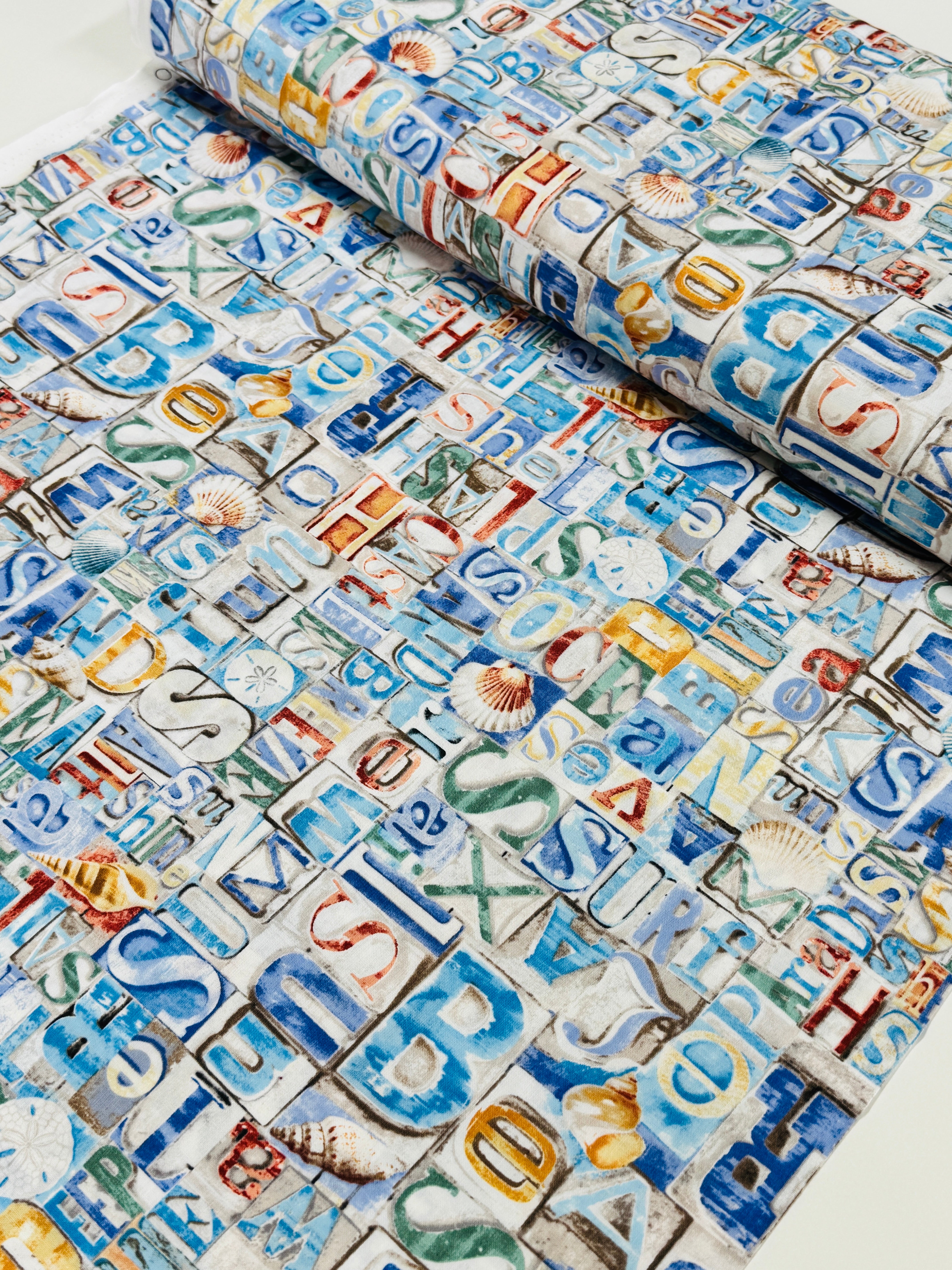 Coastal cotton fabric featuring beach-themed words, seashells, sand dollars, and nautical typography in blue, tan, and white tones.