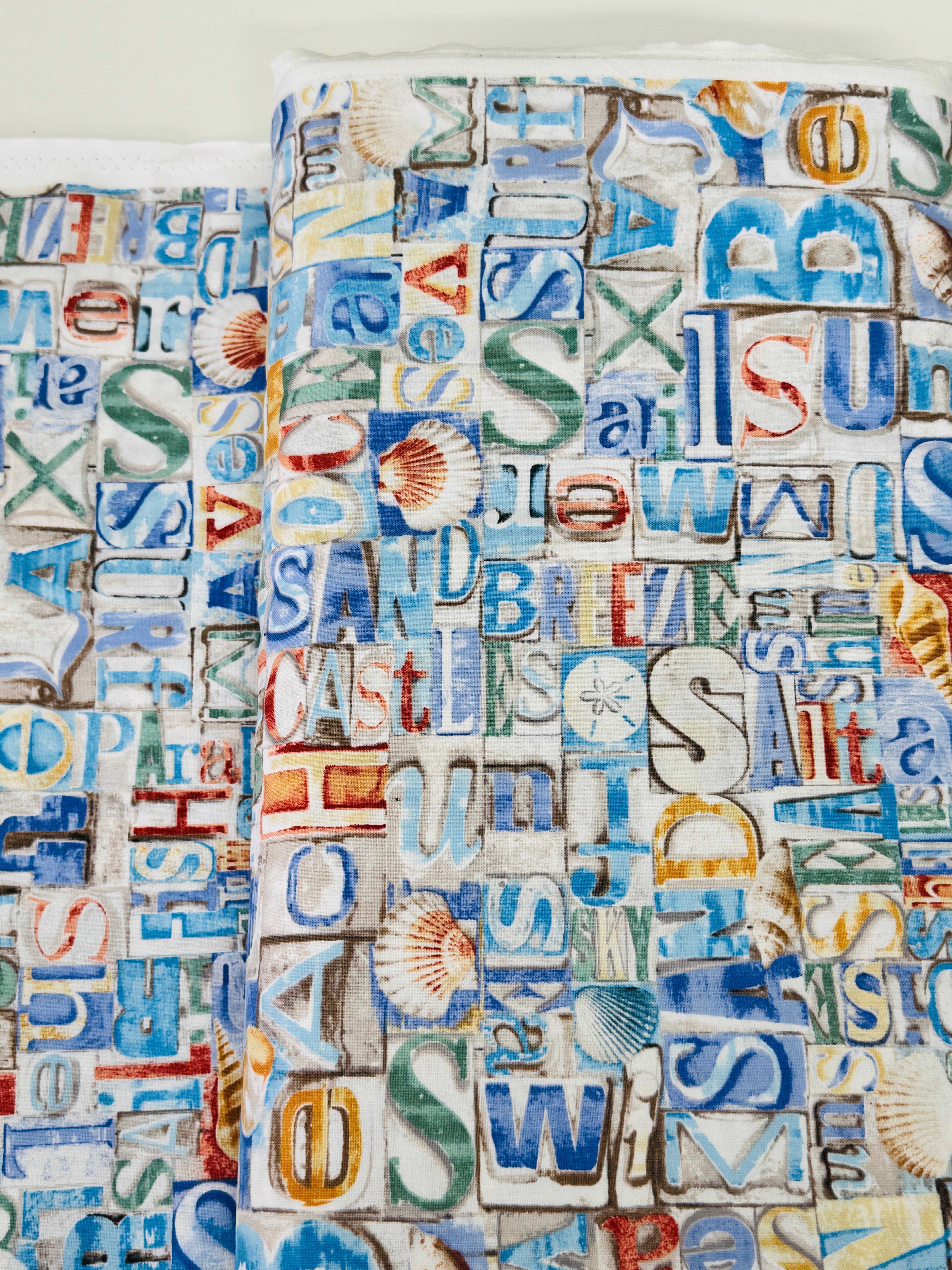 Coastal cotton fabric featuring beach-themed words, seashells, sand dollars, and nautical typography in blue, tan, and white tones.