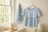 A soft blue and white gingham kitchen towel and matching blouse made from Shine – Gingham Sky Fabric by Sweetwater for Moda Fabrics, hanging near a bright window in a cozy farmhouse kitchen setting.