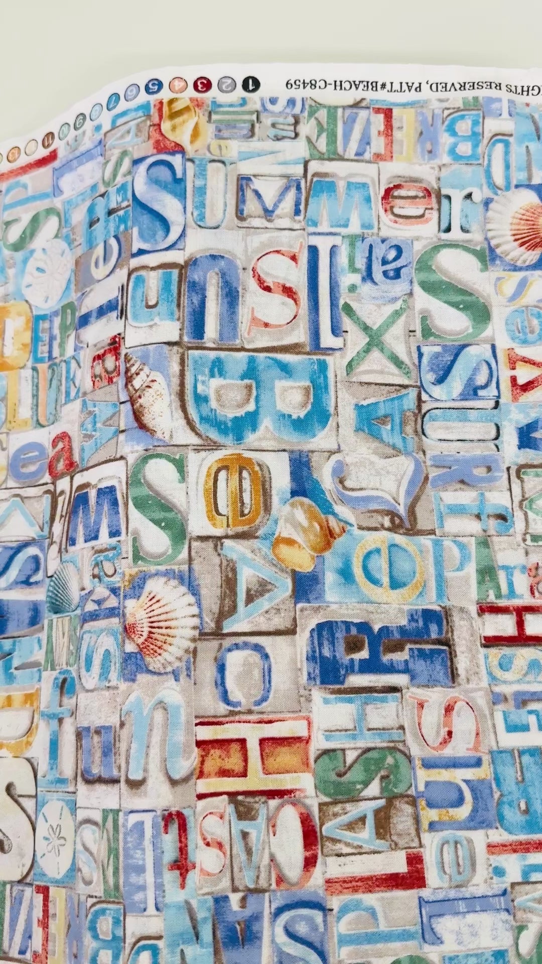 Coastal cotton fabric featuring beach-themed words, seashells, sand dollars, and nautical typography in blue, tan, and white tones.